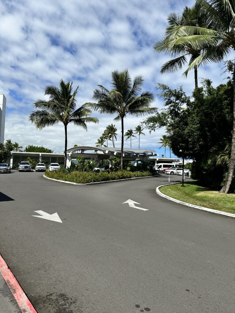 a road with palm trees and cars on the side leading to the Grand Naniloa Hotel